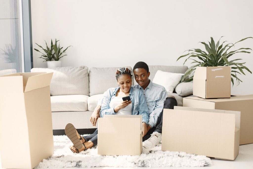 Couple sitting among moving boxes in their apartment, smiling while using a smartphone to get an instant moving quote. Photo by Pexels.