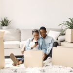 Couple sitting among moving boxes in their apartment, smiling while using a smartphone to get an instant moving quote. Photo by Pexels.