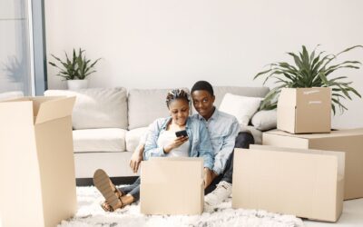 Couple sitting among moving boxes in their apartment, smiling while using a smartphone to get an instant moving quote. Photo by Pexels.