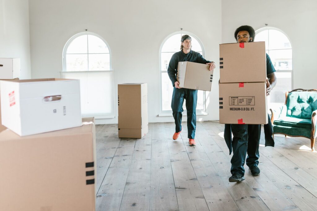 Two professional movers in uniform carrying cardboard boxes through a bright, sunlit apartment room on moving day.