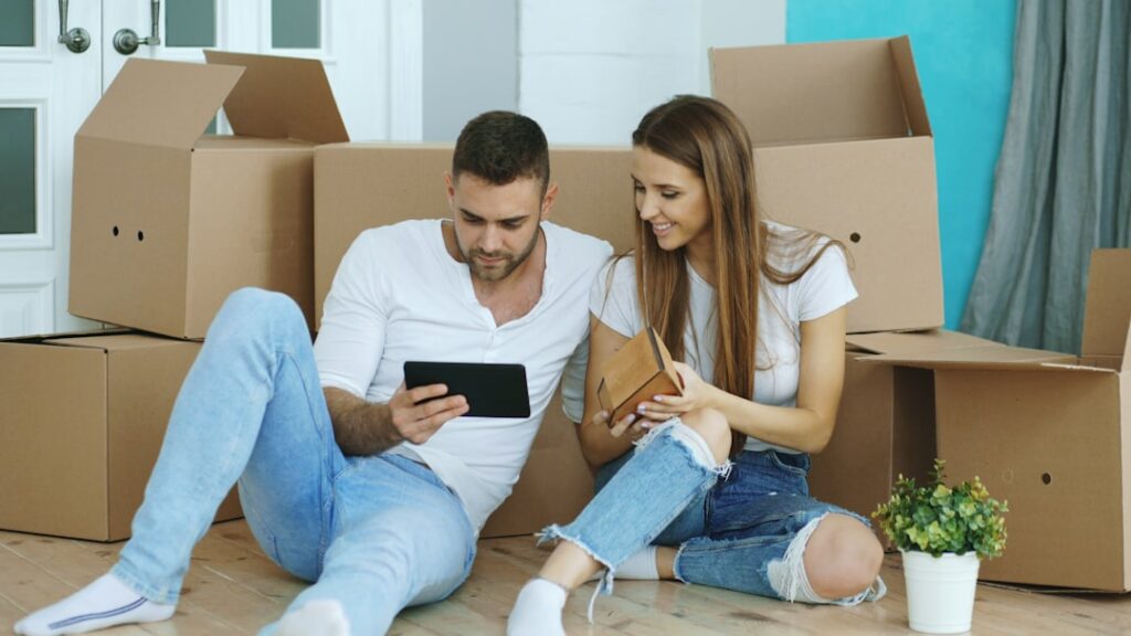 Young couple sitting on the floor surrounded by packed moving boxes reviewing their moving plan on a tablet